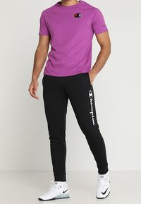 Fialové atletické tričko s krátkým rukávem a logem je zkombinováno s černými tepláky, které mají vertikální bílé logo. Celkový vzhled doplňují bílé sportovní boty.