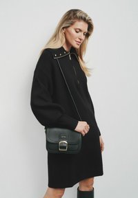 Czarna dzianinowa sukienka z wysokim kołnierzem i złotym zamkiem błyskawicznym, zestawiona z ciemnozieloną torbą na ramię z metalowym zapięciem.