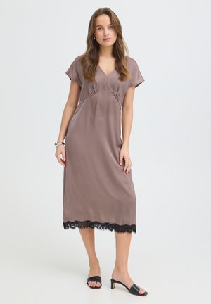 Žena stojí oblečená v taupe midi šatách s čiernym čipkovým lemom, krátkymi rukávmi, výstrihom do V a čiernymi otvorenými sandálmi na opätku na bielom pozadí.