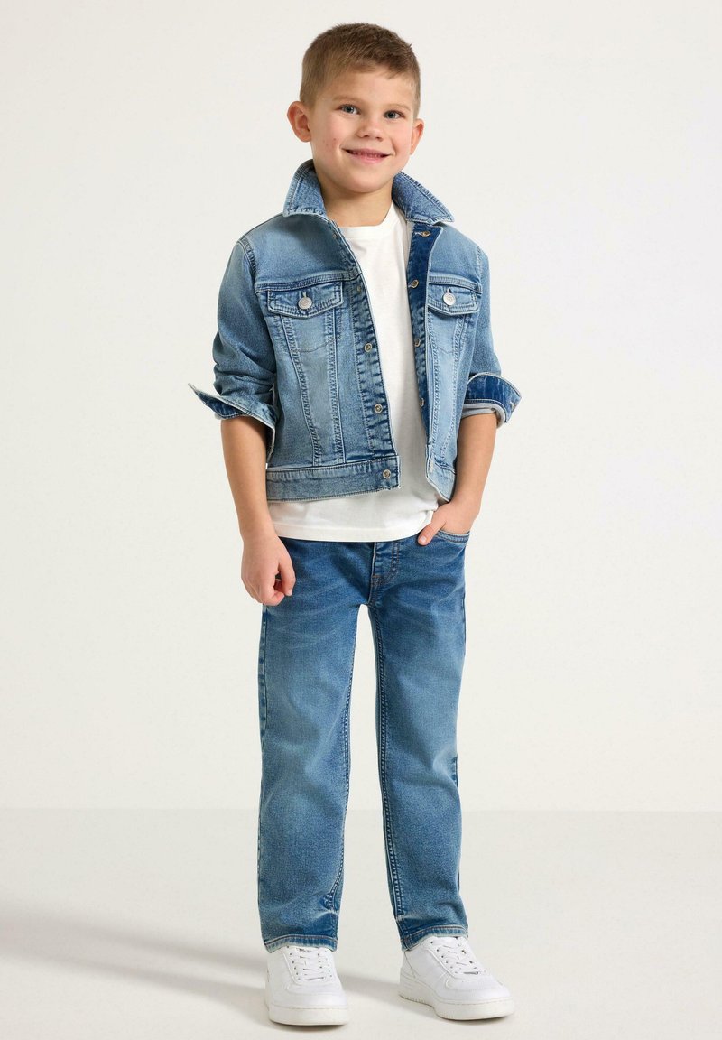 D Jeanjacke mit heller Waschung, Knopfverschluss und Brusttaschen, kombiniert mit blauen Jeans und weißen Sneakers, vor einem neutralen Hintergrund.