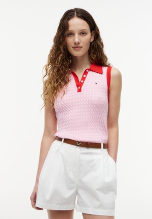 Młoda kobieta o długich kręconych włosach, ubrana w różowy dzianinowy top bez rękawów z czerwonym kołnierzem oraz białe szorty z wysokim stanem i brązowym paskiem.