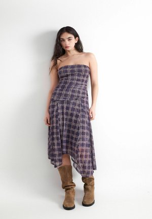 Jonge vrouw met lang donker haar, gekleed in een strapless paars geruit jurkje en slappe bruine suède laarzen tegen een witte achtergrond.
