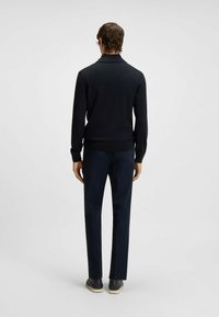 Marineblauer Strickpullover mit hohem Kragen, kombiniert mit dunklen Hosen und lässigen Schuhen. Das Material wirkt weich und hat ein tailliertes Design.