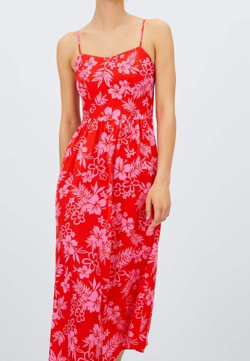 Femme portant une robe rouge sans manches avec des motifs floraux roses et de fines bretelles, debout devant un fond clair uni.