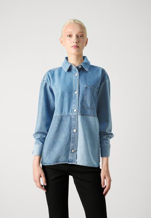 Camisa de mezclilla azul clara con frente abotonado, que presenta un bolsillo con parche y tonos contrastantes. Mangas largas con puños abotonados.