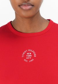 T-shirt en coton rouge avec un col rond et des manches courtes. Présente un logo brodé blanc : "TOMMY HILFIGER, NEW YORK CITY, EST. 1985."