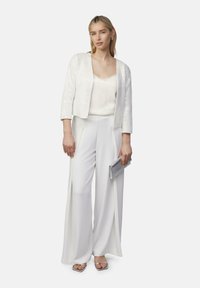 Weiße Pailletten-Cropped-Jacke über einem weißen Tanktop, kombiniert mit weiten weißen Hosen. Eine durchsichtige Clutch und metallische Sandalen runden den Look ab.