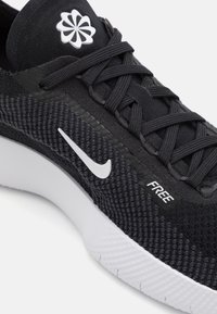 Svart atletisk sko med texturerad ovandel, vit Nike swoosh och "FREE"-etikett. Har platta snörningar och en vit, mönstrad sula.
