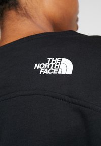 Sudadera negra con un tejido texturizado, que presenta un logo bordado en blanco de "The North Face" en la parte trasera cerca del cuello.
