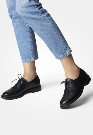 Sorte lædersko med snørebånd, glat finish, rund tå og lav hæl, kombineret med korte blå jeans, der viser et klassisk design.