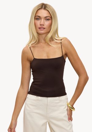 Femme aux longs cheveux blonds portant un haut à fines bretelles marron foncé, un pantalon beige clair et un bracelet en or au poignet droit.