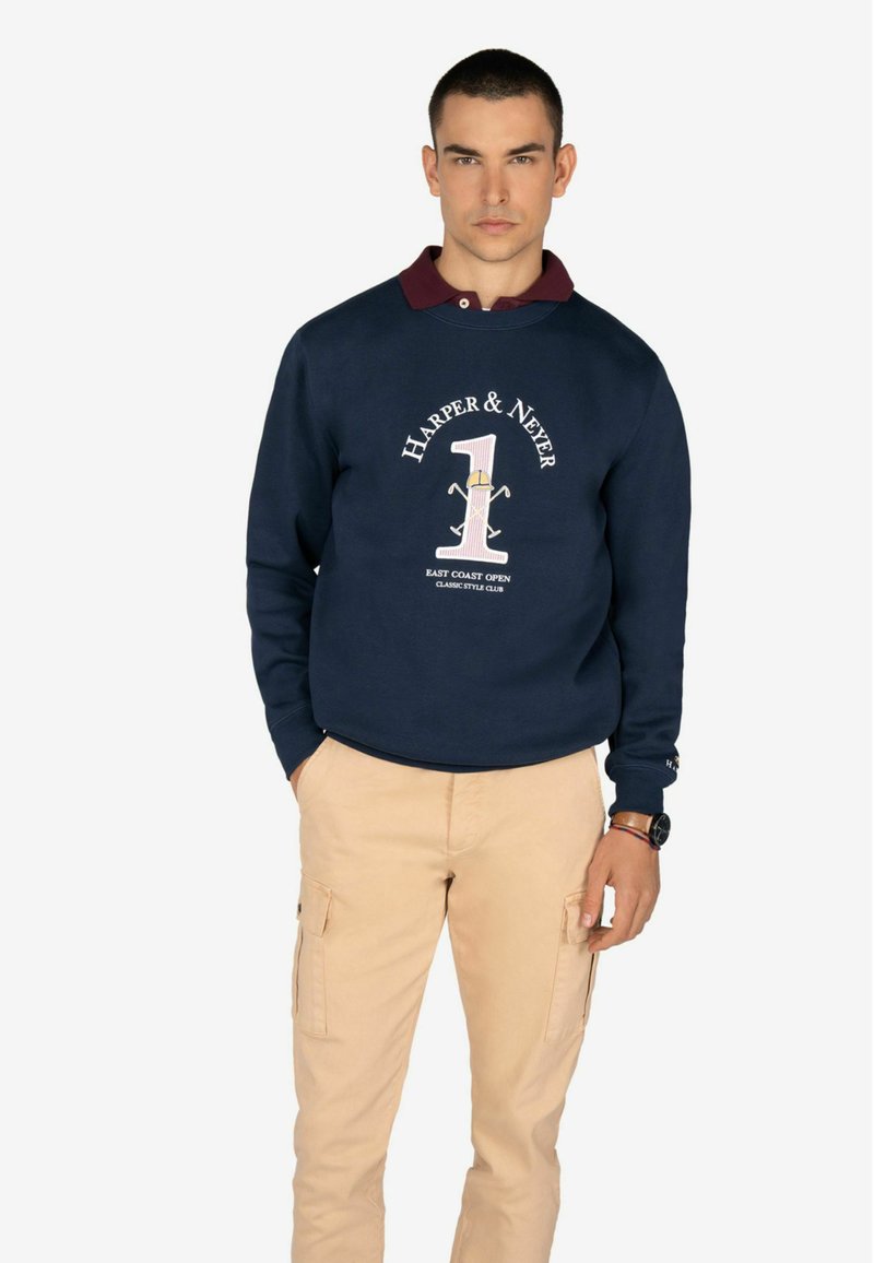 Felpa blu navy con colletto bordeaux, caratterizzata da una grande grafica che dice "HARPER & NEVER 1" con dettagli. Pantaloni cargo beige completano l'outfit.