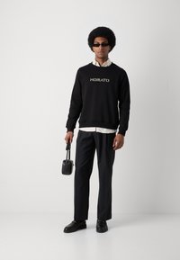 Sudadera negra con "MORATO" en fuente metálica, superpuesta a una camisa blanca. Combinada con pantalones de corte negro y loafers, sosteniendo un bolso pequeño.