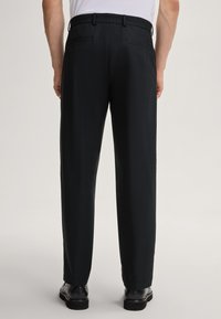 Pantalons noirs en tissu lisse, avec une coupe droite, des passants de ceinture et des poches arrière, associés à des chaussures noires.