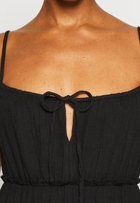 Vestido negro texturizado con tirantes finos, detalle de lazo en el escote y una cintura ceñida que crea una silueta relajada.