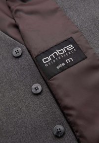 Manteau en laine gris avec des boutons noirs et une étiquette intérieure noire indiquant « ombre Quintessence taille M » sur une doublure en tissu marron lisse.