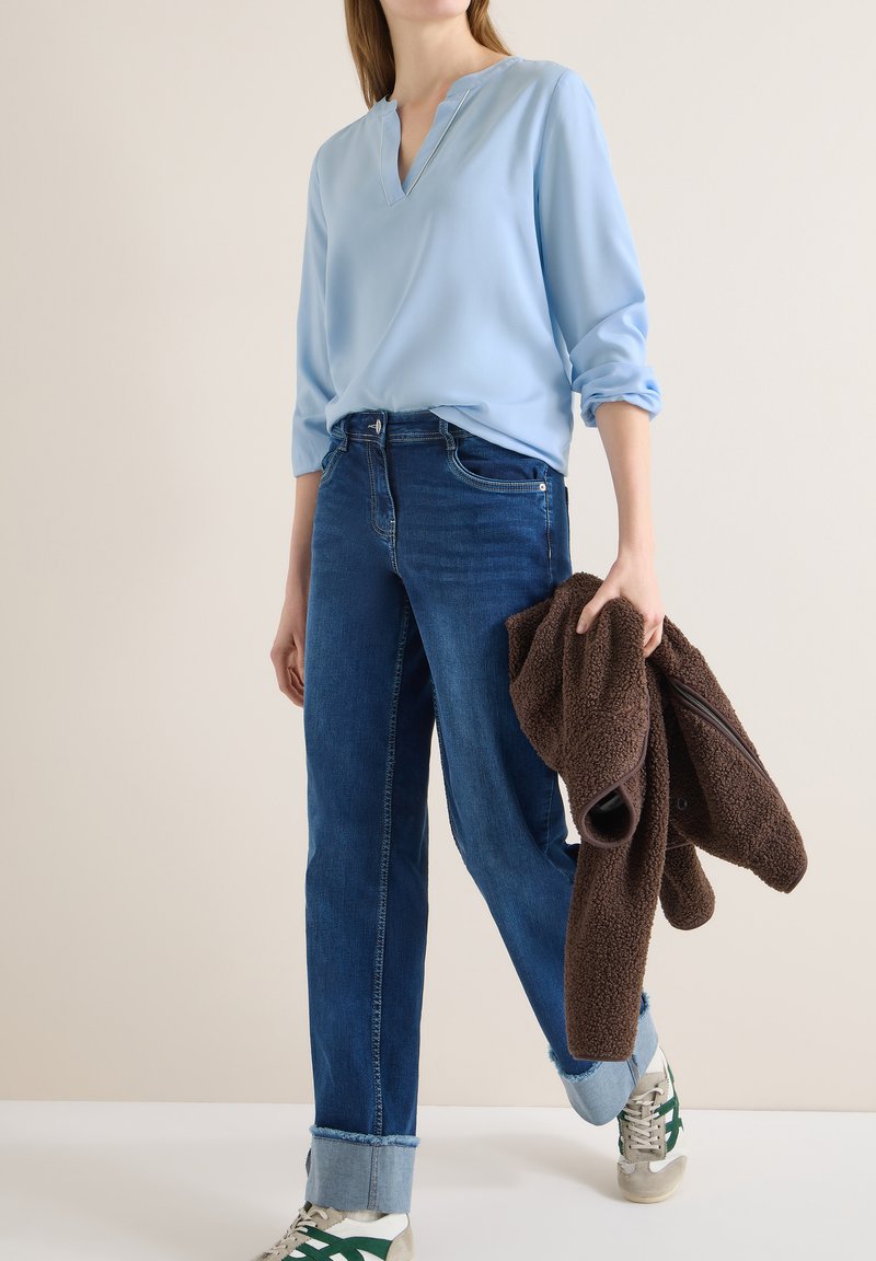 Femme portant un chemisier bleu clair et un jean foncé retroussé, tenant une veste polaire marron et portant des baskets blanches à rayures vertes.