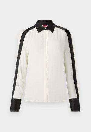 Blouse blanche avec des accents en satin noir le long des épaules et des poignets, dotée d'un col classique et de manches longues dans une texture de tissu lisse.