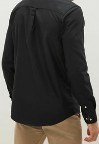 Zwart shirt met lange mouwen, een boord met knopen en een plooi aan de achterkant. Heeft een afgeronde zoom en knopen aan de manchetten.