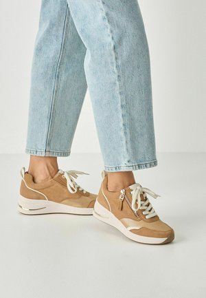 Beige suede sneakers with white accents, round toe, textured patterns, zip detail, and lace closure, paired with light blue cropped jeans.