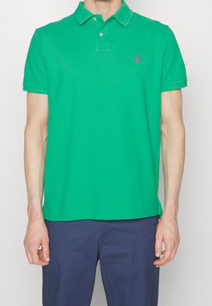 Man in een groene polo met korte mouwen en een roze geborduurd logo, gecombineerd met donkerblauwe broek, weergegeven van schouders tot heupen.