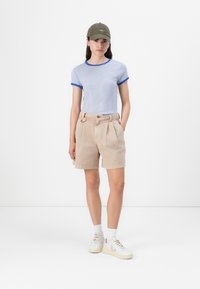 T-shirt rayé bleu et blanc avec des bordures bleues, shorts plissés beiges, et baskets blanches avec des accents gris, complétées par une casquette verte.