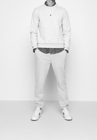 Conjunto de sudadera y pantalones de chándal en gris claro, hecho de una tela suave. Lleva un cuello redondo, puños acanalados y un logo bordado en el pecho. Zapatillas blancas.