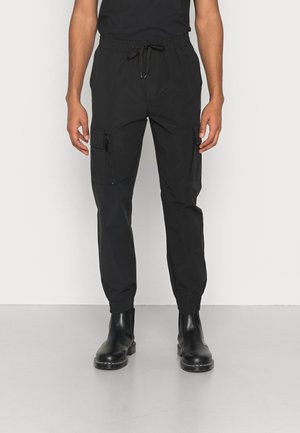 Zwart slim-fit cargobroek met elastische boorden en taille, voorzien van rits aan de voorkant en klepzakken, gedragen met zwarte enkellaarzen.