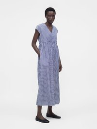 Femme debout portant une robe midi sans manches à rayures verticales bleues et blanches avec des poches et des chaussures plates noires.