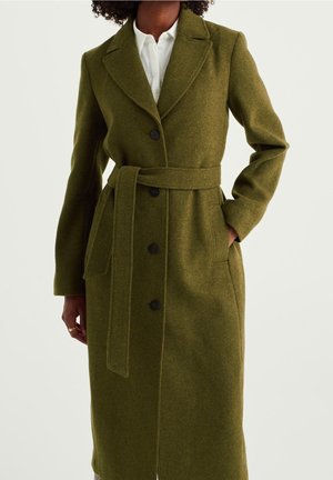 Frau trägt einen langen olivgrünen Mantel mit Gürtel und schwarzen Knöpfen über einem weißen Hemd mit Kragen, eine Hand in der Tasche.