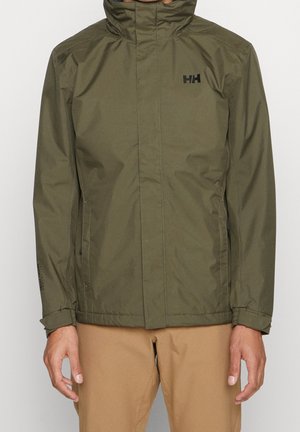 Mężczyzna w oliwkowozielonej kurtce na zamek z wysokim kołnierzem i beżowych spodniach, stojący z rękami swobodnie opuszczonymi wzdłuż ciała.