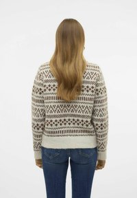 Cremfarbener Strickpullover mit einem geometrischen braunen Muster, ausgestattet mit einem runden Hals und gerippten Bündchen. Getragen mit blauen Jeans.