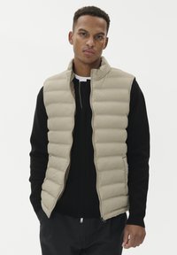 Noor mees, kes kannab beeži tikkimata käisteta vestit musta pika varrukaga särk üle, seisab tavalise valge tausta ees.