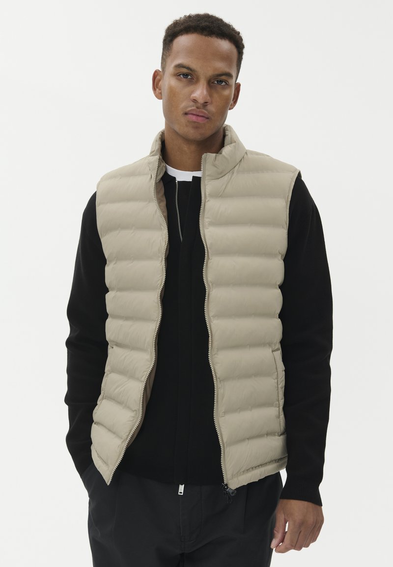 Noor mees, kes kannab beeži tikkimata käisteta vestit musta pika varrukaga särk üle, seisab tavalise valge tausta ees.