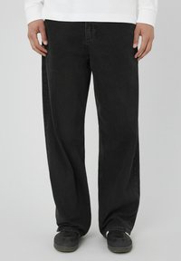 Jean noir à coupe ample, en denim, présentant une texture subtile et un design classique à cinq poches. Porté avec des chaussures sombres.
