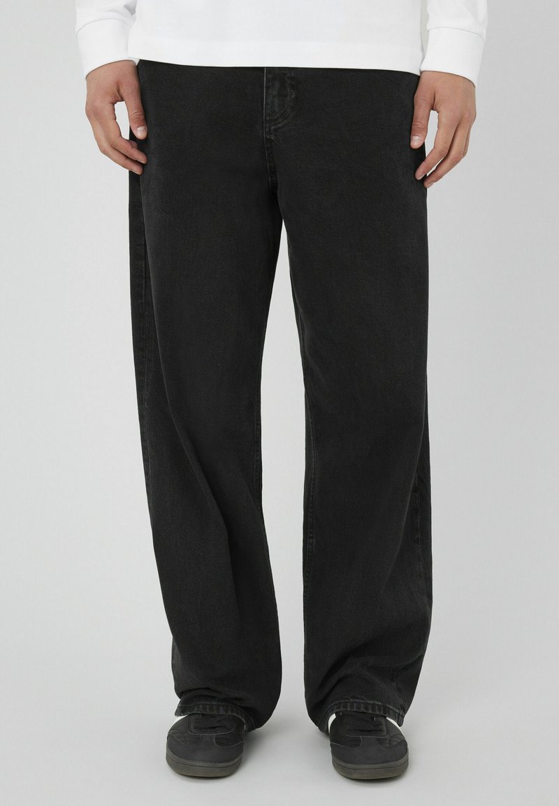 Jean noir à coupe ample, en denim, présentant une texture subtile et un design classique à cinq poches. Porté avec des chaussures sombres.