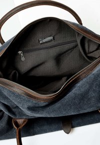 Torba denimowa z brązowymi skórzanymi akcentami, otwarta, ukazująca czarną podszewkę, kieszeń na zamek oraz etykietę wszytą w środku.