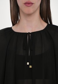 Black sheer blouse with a rounded neckline, featuring a decorative tie at the center and metallic accents at the ends of the ties.