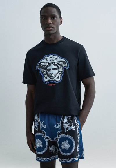 Schwarzes Baumwoll-T-Shirt mit einem Medusa-Motiv und blauen Akzenten. Kombiniert mit gemusterten blauen Shorts mit filigranen silbernen Mustern.