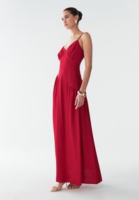 Rotes ärmelloses Maxikleid mit gerafftem Oberteil und fließendem Rock. Glatter Stoff, dezenter Glanz und Seitentaschen. Kombiniert mit neutralen Sandalen.