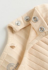 Encolure de vêtement bébé beige côtelée avec trois boutons-pression métalliques partiellement ouverts sur un fond clair.