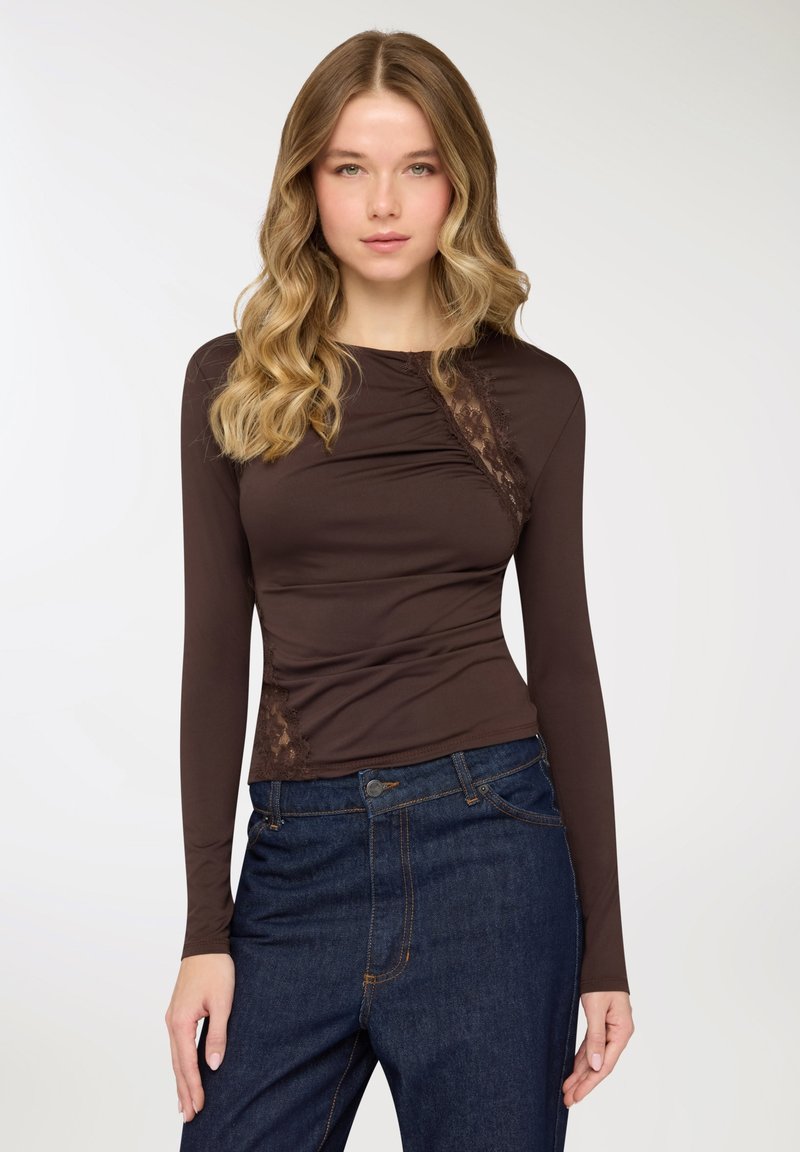 Top marrone a maniche lunghe con design aderente, caratterizzato da dettagli in pizzo sul lato sinistro e lievi arricciature lungo il busto. Indossato con jeans denim blu.