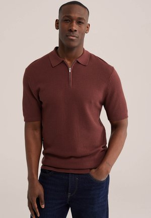 Egy férfi, aki rövid ujjú, texturált bordó polóinget visel cipzáros gallérral és sötétkék farmert, egyik kezét zsebében tartva áll.