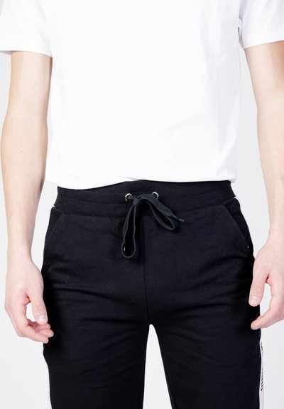 Czarne spodnie dresowe z elastycznym paskiem i sznurkiem, wyposażone w boczne kieszenie i gładką fakturę materiału. Połączone z białą koszulką.