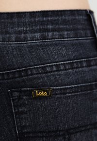Dark blue denim jeans with a smooth texture, featuring yellow logo stitching on a back pocket and subtle fading details.