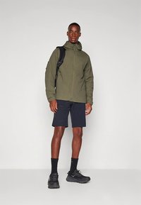 Olivengrønn jakke med glidelås, lett stoff, med hette. Kombinert med mørke shorts og svarte joggesko. Enkel, funksjonell design.