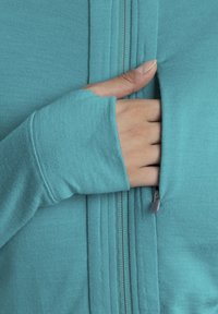 Giacca zip-up teal in tessuto morbido e testurizzato; presenta una mano che si infila in una tasca laterale, con il pollice che sbuca dalla manica lunga.