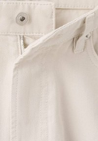 Tela de mezclilla beige con una superficie texturizada, que presenta un botón a presión plateado y una cremallera. Incluye costuras reforzadas a lo largo de las uniones.