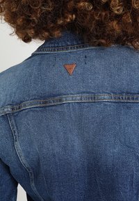 Džínová bunda ve středně modré barvě, s oranžovým prošíváním a malou hnědou trojúhelníkovou nášivkou na zadním límci. Hladký povrch.