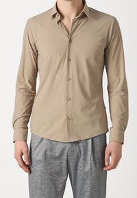 Beige långärmad skjorta med krage som knäpps ner, horisontella sömmar, slät tyg och rundad nederkant. Kombineras med texturerade grå byxor.